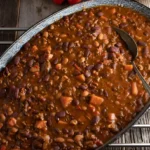 Overhead shot of smoked baked beans with chopped turkey in cast iron skillet, on rustic wooden board.